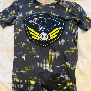 Under Armour kompressionströja med tryck - Säljer en tight kompressionst-shirt från Under Armour med stort pantertryck och gula detaljer. T-shirten har korta ärmar och är gjord i syntetmaterial, perfekt för träning eller sport.
