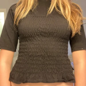 Svart smockad topp med halvlånga ärmar - Svart topp med smockad midja och halvlånga ärmar. Toppen har en lätt volangkant nertill och en hög hals. Perfekt för en trendig och bekväm look.