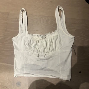 Vit linnetopp med rosett från Gina Tricot - Säljer en söt vit linnetopp från Gina Tricot. Toppen har smala axelband, rynkade detaljer och en liten rosett framtill. Perfekt för varma dagar och enkel att matcha med olika outfits.