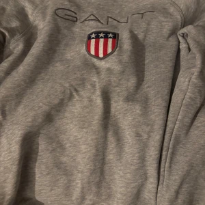 Grå sweatshirt från GANT med broderad logga - Säljer en grå sweatshirt från GANT med broderad logga och sköld på bröstet. Tröjan har rund halsringning och klassisk passform. Perfekt för en avslappnad och stilren look. Den är i smal barn storlek.