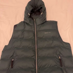 Mörkgrön dunväst med huva från GANT - Snygg mörkgrön dunväst från GANT med huva och dragkedja framtill. Västen har två fickor med dragkedja och är perfekt för lager-på-lager. Klassisk logga på bröstet och quiltad design.