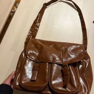 Brun axelväska i skinn med fickor - Säljer en brun axelväska i skinn med två stora fickor framtill och justerbar axelrem. Väskan har lock med spännen och magnetknappar samt dragkedja upptill. Perfekt för dig som vill ha plats med mycket och gillar klassisk stil.