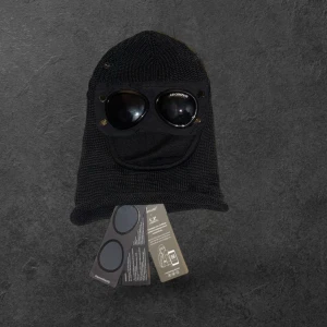 Svart goggle-mössa från C.P. Company - Säljer en svart stickad goggle-mössa från C.P. Company. Mössan har inbyggda svarta glasögon framtill och är tillverkad i ett grovstickat material. Perfekt för dig som vill sticka ut med en unik accessoar.