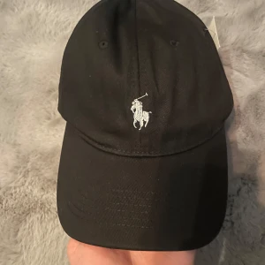 Svart keps från Polo Ralph Lauren - Svart keps med klassisk Polo Ralph Lauren-logga broderad framtill och texten POLO baktill. Justerbart spänne i metall bak för perfekt passform. Snygg och stilren accessoar som passar till det mesta.