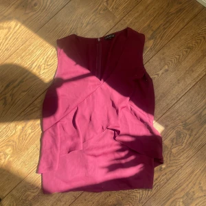 Vinröd ärmlös klänning med volang från Vero Moda - Säljer en vinröd ärmlös klänning från Vero Moda med v-ringning och dekorativ volang framtill. Klänningen har en elegant och stilren design, perfekt för dig som vill sticka ut med färg. Dragkedja i ryggen för enkel påklädning.