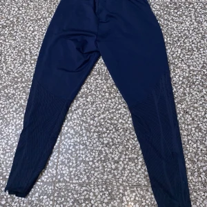 Mörkblå träningsbyxor från Nike - Säljer ett par mörkblå träningsbyxor från Nike med smal passform och elastisk midja med dragsko. Byxorna har diskret mönster på benen och en vit Nike-logga på vänster sida. Perfekta för gymmet eller löprundan.