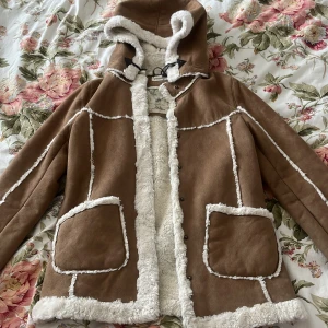 Brun teddyjacka med huva från H&M Young - Säljer en gosig brun teddyjacka från H&M Young med vit fuskpäls på insidan, huva och stora fickor framtill. Jackan är från från Young men kan användas perfekt för kvinnor i storlek S. Jackan har dekorativa sömmar och stängs med knappar. Samt har jackan en luva med päls i. Perfekt för kyliga dagar!