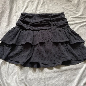 Svart volangkjol - Supersöt svart kjol med volanger och strukturmönster i tyget. Kjolen har resår i midjan och är kort i modellen, perfekt för en trendig look. Passar till många olika stilar och är lätt att matcha med olika toppar.