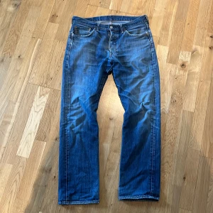 Levis 501 blå jeans - Klassiska blå Levis 501 jeans med raka ben och normal passform. Jeansen har fem fickor, knappgylf och tydliga Levis-detaljer som röd etikett och läderpatch bak i midjan. Perfekta för en tidlös och avslappnad stil. Snygga slitningar med litet hål vid knät. Kan skicka med lagnings patch för den som vill ha, bara att stryka på. Strl 33/32