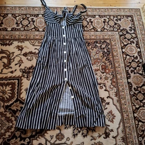 Svartvit randig klänning med knappar - Säljer en svartvit randig klänning med smala axelband och knyt fram. Klänningen har knappar längs hela framsidan och är i en längre modell. Perfekt för varma dagar och har en luftig passform.