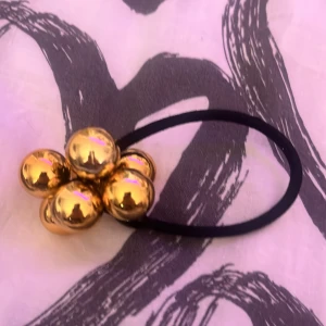 Guldig hårsnodd med klot - Säljer en svart hårsnodd med fem stora, blanka guldfärgade klot som dekoration. Perfekt för att ge en extra touch till din frisyr. Enkel att använda och snygg detalj i håret.