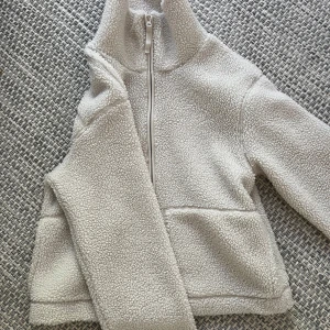 Vit teddyjacka med dragkedja - Mysig vit teddyjacka med hög krage och hel dragkedja framtill. Jackan har två stora fickor och är tillverkad i ett fluffigt, fleece-liknande material. 