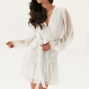 Studentklänning/ vit omlottklänning - Superfin vit kortklänning med omlott och knyt i midjan. Klänningen har långa ballongärmar i transparent tyg och volangdetalj nedtill. Perfekt för sommarens tillfällen och student. Oanvänd! Ordinarie pris 699kr