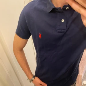 Mörkblå Piké från Polo Ralph Lauren - Säljer en klassisk mörkblå pikétröja från Ralph Lauren med röd broderad logga på bröstet. Tröjan har korta ärmar, krage och knappar vid halsen. Perfekt för en stilren och avslappnad look. Storlek Large Custom Fit. Snabb Leverans!