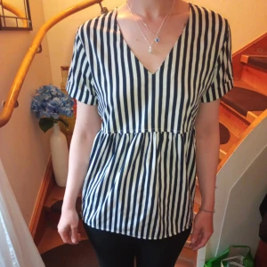 Randig blus med v-ringning från ICHI - Säljer en snygg blus från ICHI med marinblå och vita vertikala ränder. Blusen har v-ringning framtill, korta ärmar och en öppen detalj i ryggen. Perfekt för dig som gillar en stilren och trendig look.