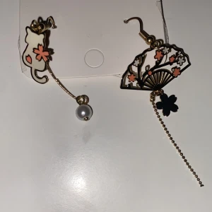 Japanska Sakura örhängen med katt och solfjäder INGA PLUPPAR - Säljer ett par unika örhängen i guld med motiv av en katt och en solfjäder, dekorerade med rosa och svarta blommor samt en vit pärla. Ena örhänget har en kedja med svart blomdetalj. Perfekt för dig som gillar söta accessoarer med japansk touch.