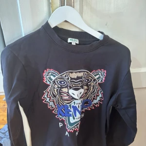Svart sweatshirt med tigerbroderi från Kenzo - Svart sweatshirt från Kenzo med stort broderat tigerhuvud och text på framsidan. Tröjan har rund halsringning och långa ärmar. Perfekt för dig som vill ha en statement-piece i garderoben.