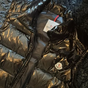Svart glansig dunväst från Moncler - Snygg svart glansig dunväst från Moncler med dragkedja och logotyp på bröstet. Väldigt sparsamt använd. Passar i M och L