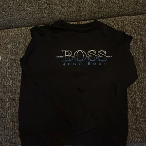 Svart sweatshirt från Hugo Boss - Säljer en svart sweatshirt från Hugo Boss med stor vit och blå logga framtill. Tröjan har rund halsringning och långa ärmar. Perfekt för en stilren och avslappnad look.