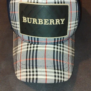 Rutig keps från Burberry - Snygg keps från Burberry med klassiskt rutigt mönster i blått, vitt, svart och rött. Framsidan har en svart patch med Burberry-logga. Baksidan är i mesh-tyg för extra ventilation. Justerbar passform.