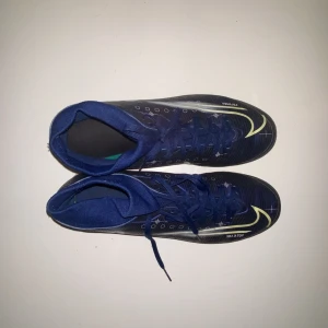 Blå Nike fotbollsskor med snörning - Säljer ett par blå Nike fotbollsskor med snörning och högt skaft. Skorna har vita och gula detaljer samt rund tå och platt sula. Perfekta för inomhusfotboll eller träning.