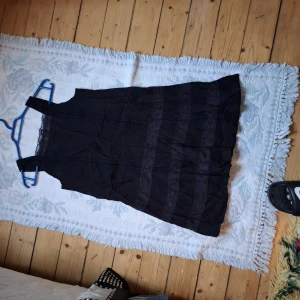 Svart ärmlös klänning med spetsdetaljer - Svart ärmlös klänning med breda axelband och fina spetsdetaljer längs kjolen. Klänningen har en lätt utställd form och är perfekt för varma dagar. Enkel och stilren design som passar till många tillfällen.