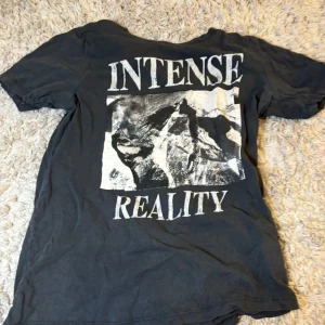 Svart t-shirt med tryck 'Intense Reality' - Cool svart t-shirt med stort vitt tryck på bröstet där det står 'Intense Reality' och ett grafiskt motiv i mitten. Perfekt för dig som gillar statement-plagg och streetstyle.