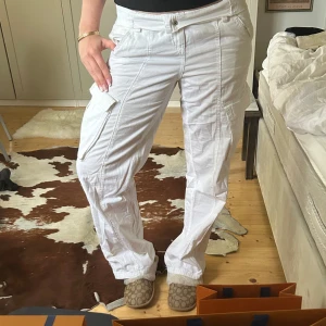 Vita cargopants med fickor - Säljer ett par vita cargopants med flera fickor och dragkedjedetaljer. Byxorna har en lös passform och raka ben, perfekt för en avslappnad stil. Stängs med knapp och dragkedja framtill.