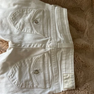 Vita jeansbyxor från True Religion - Säljer ett par vita jeansbyxor från True Religion med klassiska bakfickor och silverfärgade knappar. Byxorna har normal passform och är tillverkade i bomull. Perfekta för dig som vill sticka ut med en clean look.