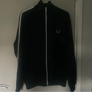 Svart track jacket från Fred Perry - Säljer en svart track jacket från Fred Perry med vita ränder längs ärmarna och vit dragkedja. Jackan har hög krage och den klassiska Fred Perry-loggan broderad på bröstet. Perfekt för en sportig och stilren look. Den är i storlek S och har en bra passform! Priset kan diskuteras bara hör av dig så kan vi se vad vi kan göra😊