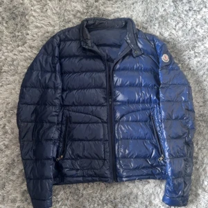 Blå pufferjacka från Moncler acorus - Säljer en blå pufferjacka från Moncler med klassisk logotyp på ärmen. Jackan har dragkedja framtill och två snedställda fickor med dragkedja. Lätt glansigt material och quiltad design ger en modern look.
