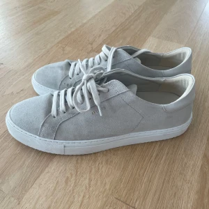 Axel Arigato Ljusgråa sneakers med snörning - Säljer ett par stilrena ljusgråa Axel Arigato sneakers i mocka med vit platt sula och rund tå. Skorna har klassisk snörning och en minimalistisk design som passar till det mesta. Perfekta för dig som gillar en clean look.