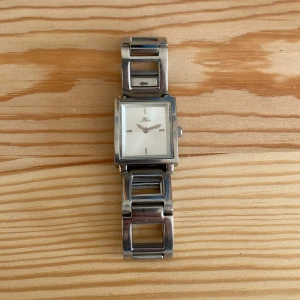 Silverfärgad klocka från Lacoste - Clean klocka från Lacoste med fyrkantig urtavla och länkarmband i silverfärgat rostfritt stål. Urtavlan är vit med minimalistiska markeringar och Lacoste-logga. Fungerar!