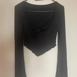 Svart långärmad croppad topp - Säljer en svart långärmad topp med v-ringning och twistad detalj framtill. Toppen är croppad med asymmetrisk nederkant och har en tight passform. Perfekt att styla med höga jeans eller kjol.🖤