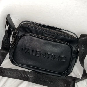 Svart axelväska från Valentino - Svart Valentino axelremsväska med justerbar axelrem med Valentino logotyp. Väskan har ett fack med dragkedja upptill och ett mindre fack framtill med Valentino logotyp.  Köpt 2022, använd ett antal gånger.  Inga ytliga skador/repor. Originalpris: ca 1300kr