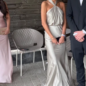 Champagnefärgad långklänning med halterneck - Säljer en elegant champagnefärgad långklänning med halterneck och draperad framsida. Klänningen har en slits och är axelbandslös med en snygg omlottkänsla. Perfekt för speciella tillfällen.