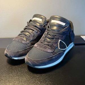 Mörkblå sneakers från Philippe Model - Snygga mörkblå sneakers från Philippe Model med ovandel i mocka och textil, samt detaljer i skinn. Skorna har snörning och vit sula med blå inslag. Perfekta för en stilren och sportig look.