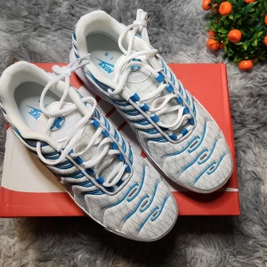 Nike Air Max Plus vita och blå sneakers - Nike Air Max Plus sneakers i vitt med blå detaljer och svarta linjer. Skorna har tydlig Tn-logo på hälen, vit platt sula och rund tå. Ovandelen är i syntetmaterial med blå Nike-logga på plösen och blå accenter längs snörningen.