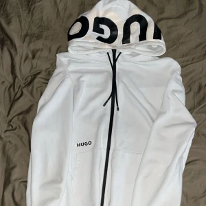 Hugo zipper - Vit hoodie från Hugo med svart logotyptryck på huvan och svart dragkedja. Klassisk passform med fickor framtill och justerbar huva med snörning. Perfekt för en stilren och avslappnad look. Väldigt sällsynt model säljs ej längre