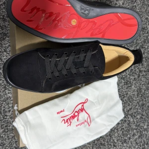 Svarta sneakers från Christian Louboutin - Säljer ett par svarta sneakers från Christian Louboutin med klassisk röd sula och diskret broderad logga på hälen. Skorna har snörning och är tillverkade i mocka med svarta detaljer och rund tå.