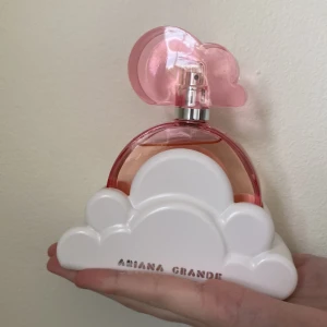 Ariana Grande Cloud Pink Eau de Parfum - En söt parfym från Ariana Grande med en rund flaska i rosa glas och ett lock format som ett rosa moln. Flaskan står i en vit molnformad bas med loggan framtill. Doften är fruktig och feminin, perfekt för dig som gillar lekfulla och trendiga parfymer.