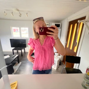 Rosa blus med volangdetaljer - Säljer en söt rosa blus med volangdetaljer vid axlarna och v-ringning. Blusen har korta ärmar och en lös passform. Toppen är från Gina och förhoppningsvis din nya sommardröm!!!!