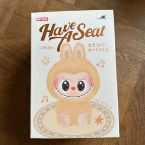 Have A Seat LABUBU - Pop Mart samlarfigurbox - Säljer en oöppnad box med samlarfigurer från Pop Mart-serien 'Have A Seat' med LABUBU-karaktärer. Förpackningen är dekorerad med söta illustrationer av olika figurer i pastellfärger och har en stilren, ljus design. Perfekt för dig som gillar samlarleksaker och kawaii-estetik.