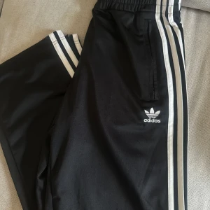Svarta träningsbyxor från Adidas med vita ränder - Klassiska svarta träningsbyxor från Adidas med de ikoniska tre vita ränderna längs sidorna och Adidas-logga på benet. Byxorna har resår i midjan och raka ben. Perfekta för sport eller chill.