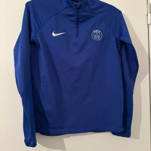 Blå långärmad träningströja från Nike PSG - Säljer en blå långärmad träningströja från Nike med PSG-logga på bröstet. Tröjan har dragkedja vid halsen och är perfekt för fotbollsträning. Snygg sportig design med diskreta mönster på ärmarna. Den säljs ej längre också, Byxorna finns också