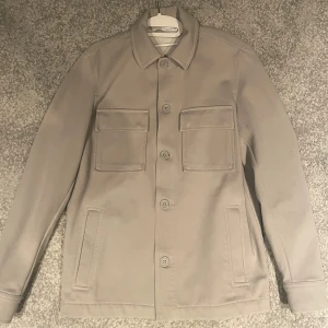 Overshirt - Stilren beige overshirt från Zara med klassisk krage och stora bröstfickor med lock. Jackan har knäppning framtill och raka ärmar. Perfekt för lager-på-lager och enkel att matcha med olika outfits.
