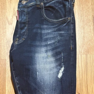Mörkblå slitna jeans från Dsquared2 - Säljer ett par mörkblå jeans från Dsquared2 med slitningar och distressed detaljer på benen. Klassisk femficksmodell med raka ben och snygga kontrastsömmar. Perfekta för en cool och avslappnad stil.
