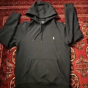 Svart hoodie från Polo Ralph Lauren - Säljer en svart hoodie från Polo Ralph Lauren med klassisk logga broderad på bröstet. Tröjan har huva med dragsko och en stor magficka framtill. Perfekt för en avslappnad och stilren look. Ordinära pris (2,500kr)