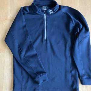 Mörkblå långärmad tröja från FootJoy - Säljer en mörkblå långärmad tröja från FootJoy med halv dragkedja framtill och hög krage. Perfekt för kyligare dagar på golfbanan eller till vardags. Tröjan har en sportig look och är tillverkad i ett mjukt syntetmaterial.