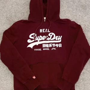 Vinröd hoodie från Superdry - Säljer en vinröd hoodie från Superdry med vit logga och text framtill. Tröjan går ej att få tag på och är väldigt snygg, priset går att diskutera vid snabb och smidig affär😄🌟
